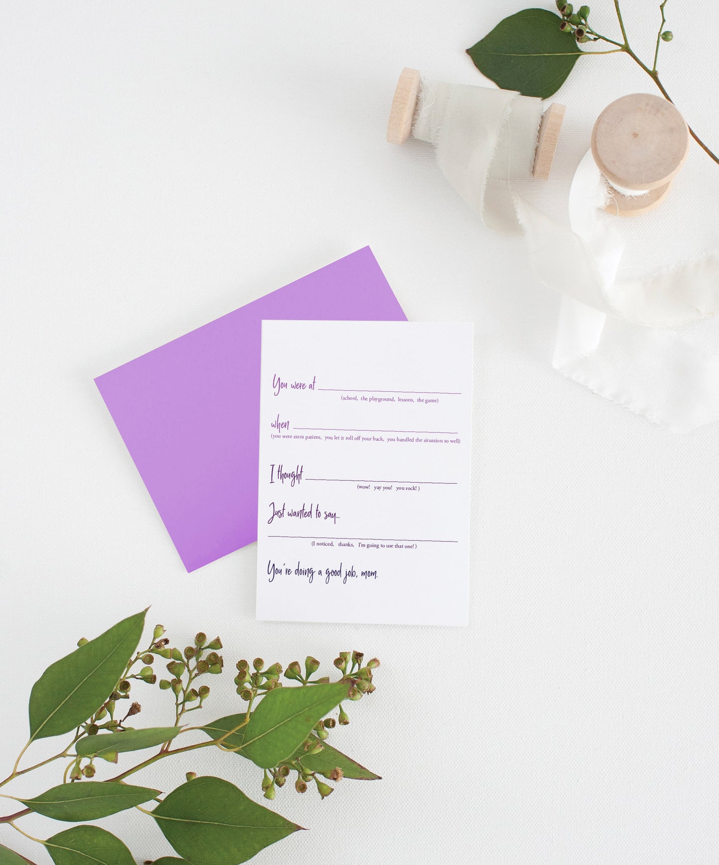 Greeting card with colorful envelope laying on a white surface with green leaves and cotton ribbon wound on wooden spools.