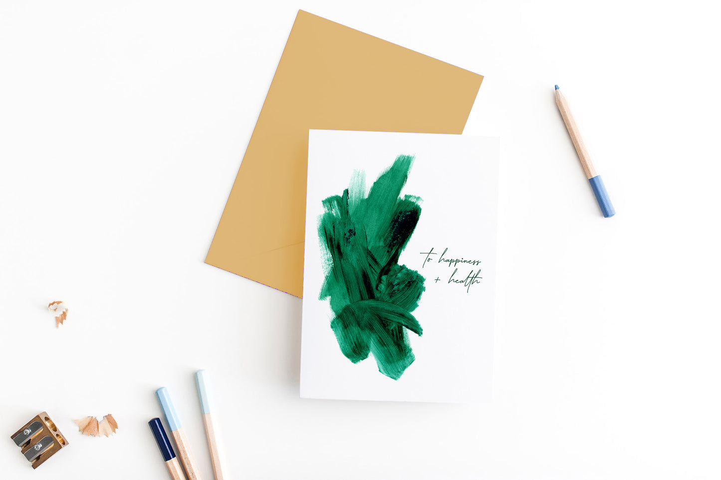 Greeting card, seen from above, with a colored, square flap envelope, pencils, pencil sharpener and shavings on a white background.