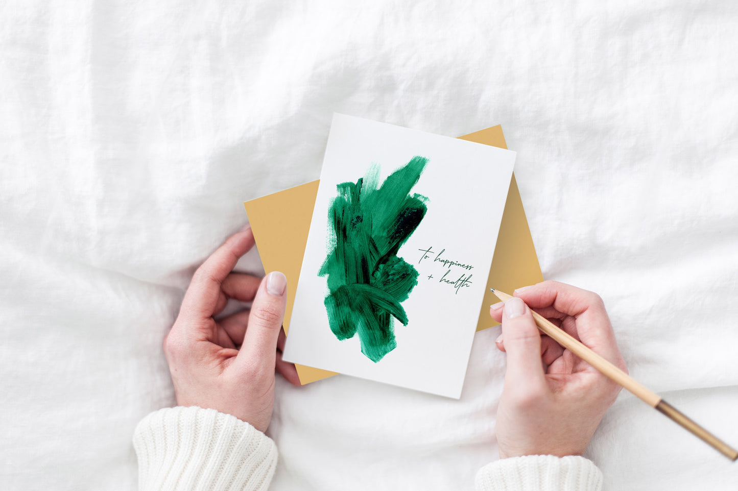 Greeting card and colorful envelope, seen from above, held by two hands along with a pencil, ready to write, against a white comforter.