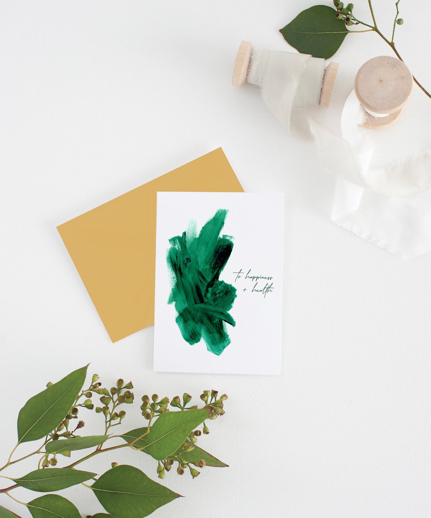 Greeting card with colorful envelope laying on a white surface with green leaves and cotton ribbon wound on wooden spools.