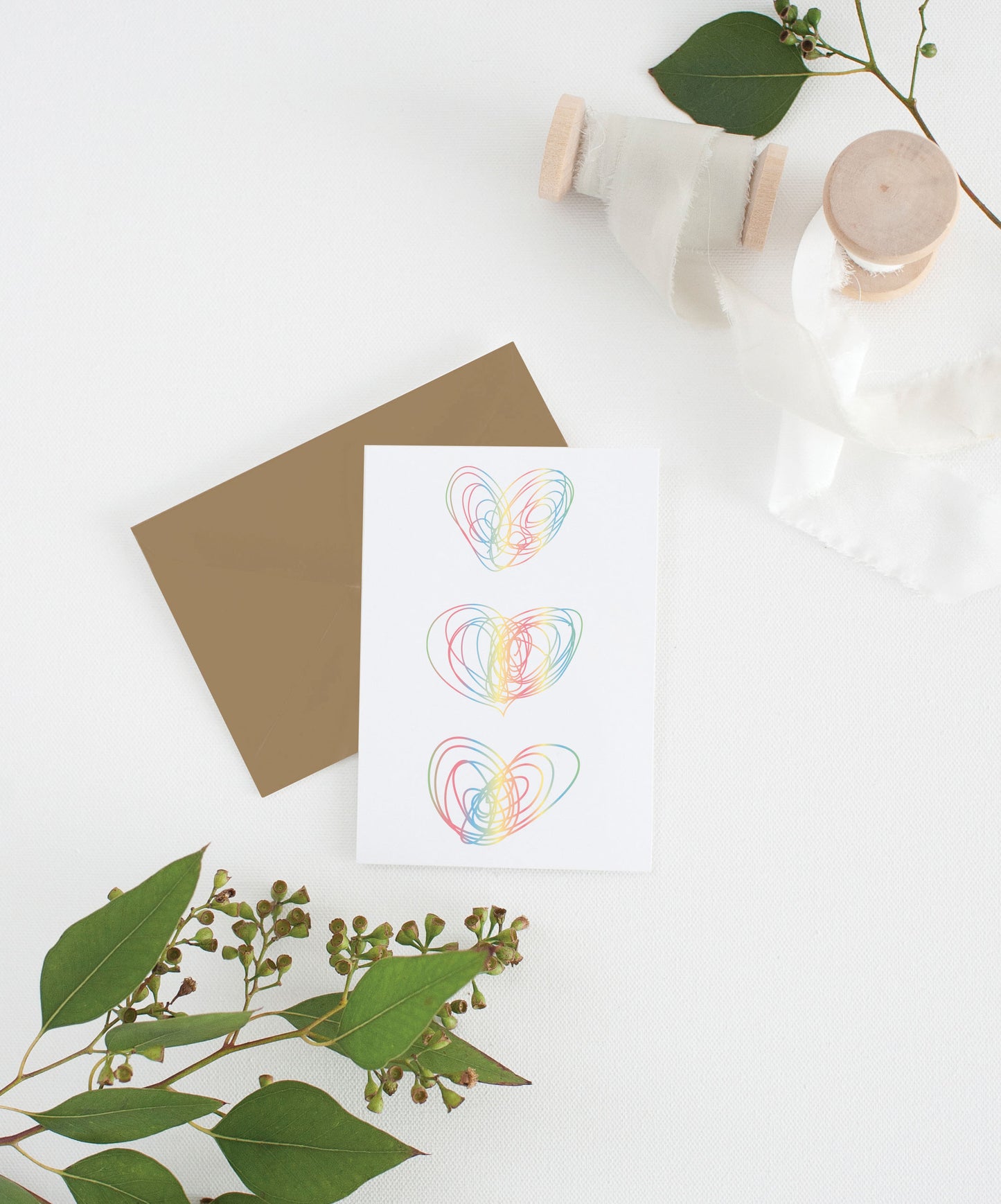 Greeting card with colorful envelope laying on a white surface with green leaves and cotton ribbon wound on wooden spools.