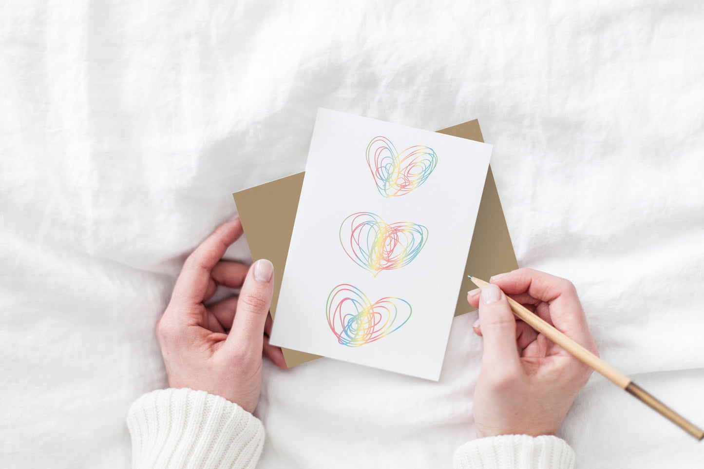 Greeting card and colorful envelope, seen from above, held by two hands along with a pencil, ready to write, against a white comforter.