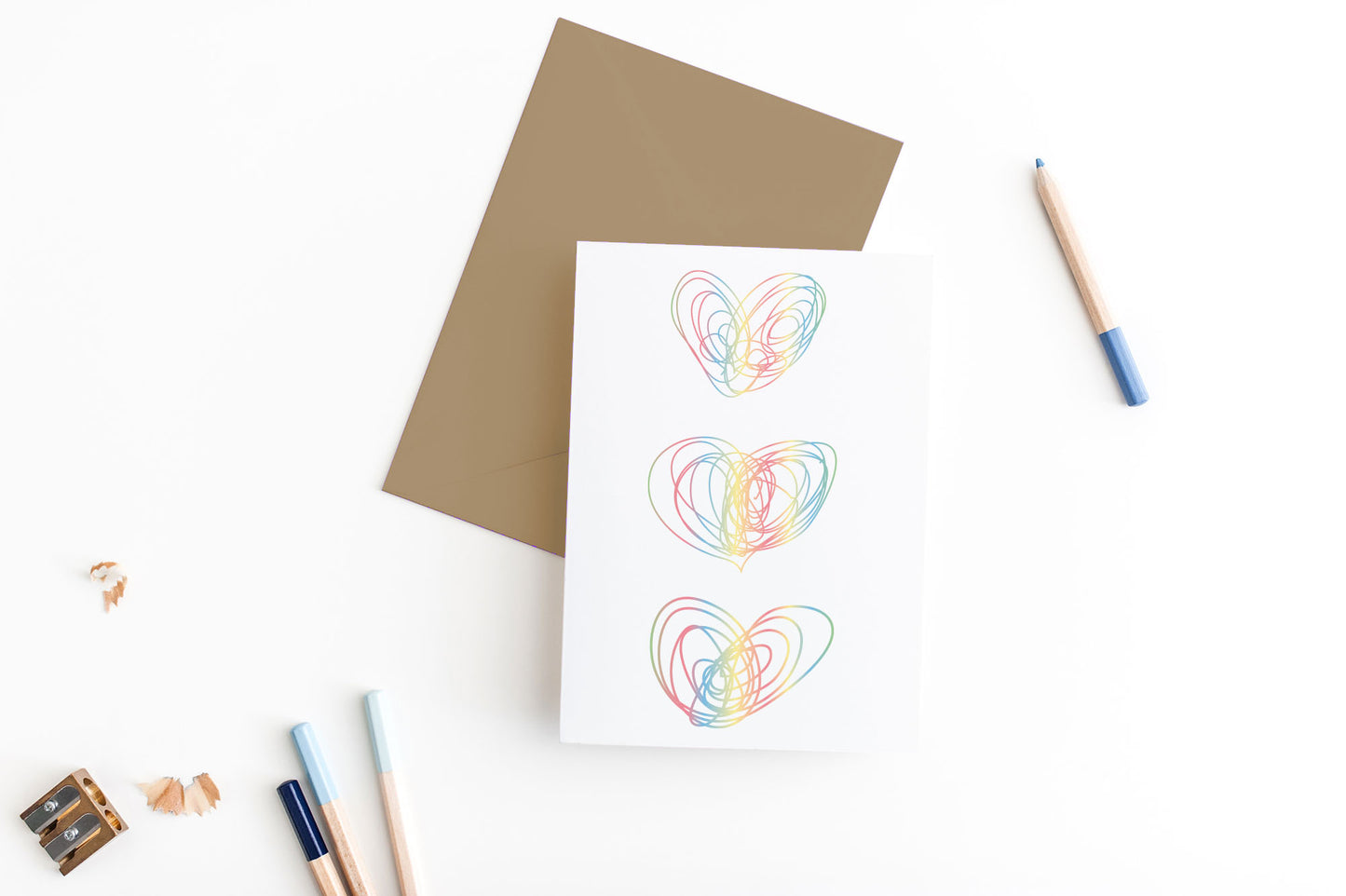 Greeting card, seen from above, with a colored, square flap envelope, pencils, pencil sharpener and shavings on a white background.