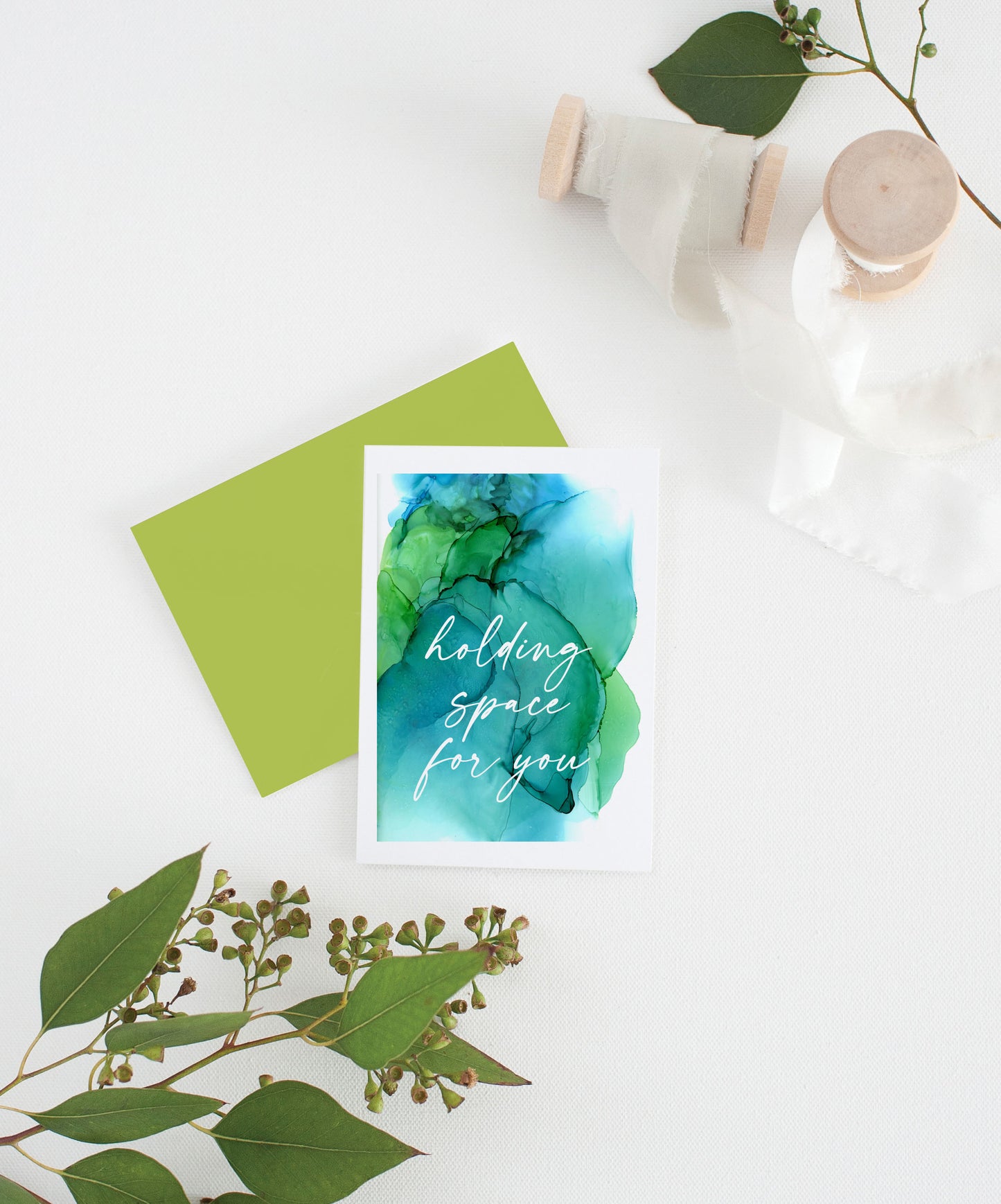 Greeting card with colorful envelope laying on a white surface with green leaves and cotton ribbon wound on wooden spools.