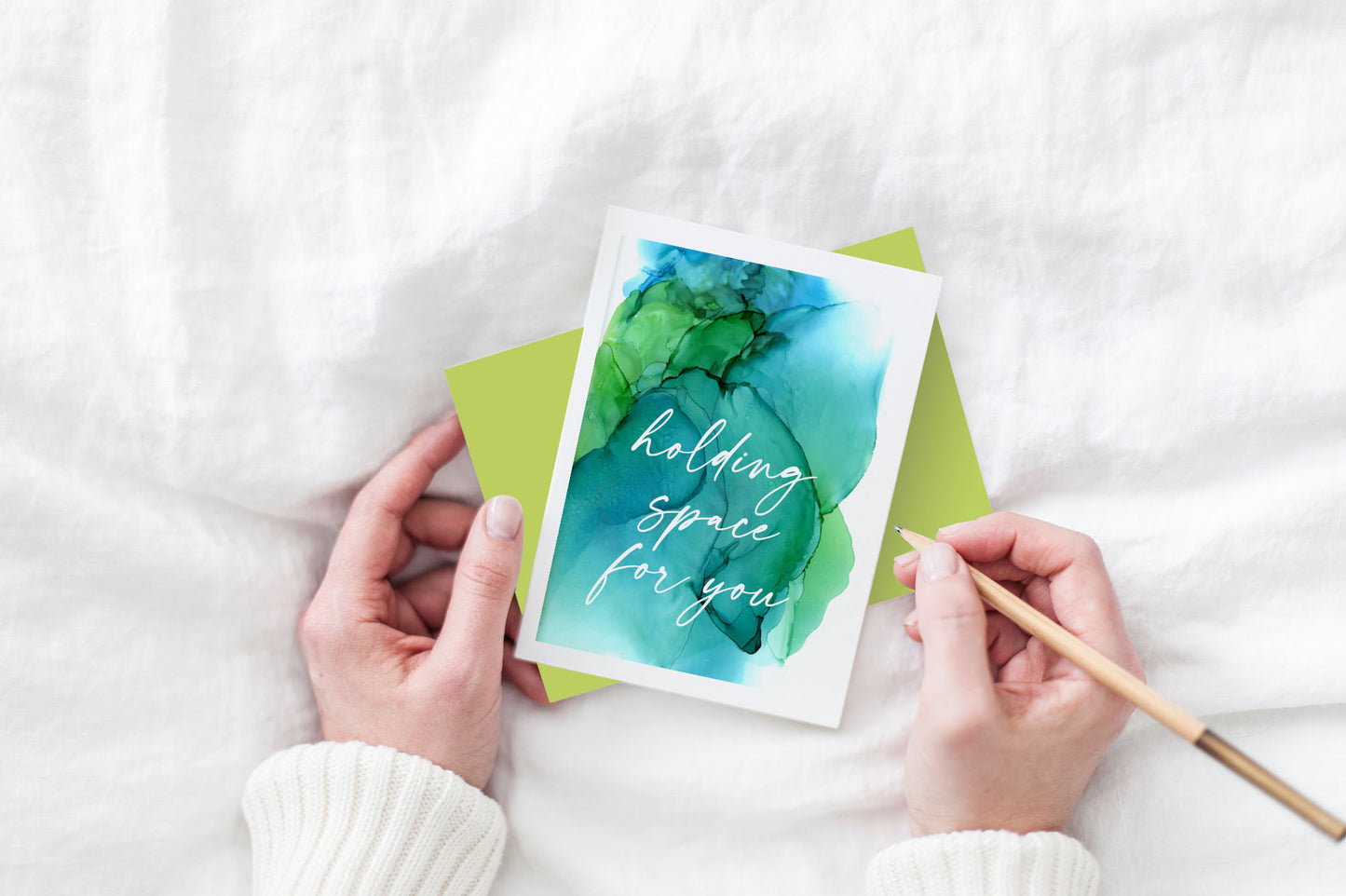Greeting card and colorful envelope, seen from above, held by two hands along with a pencil, ready to write, against a white comforter.
