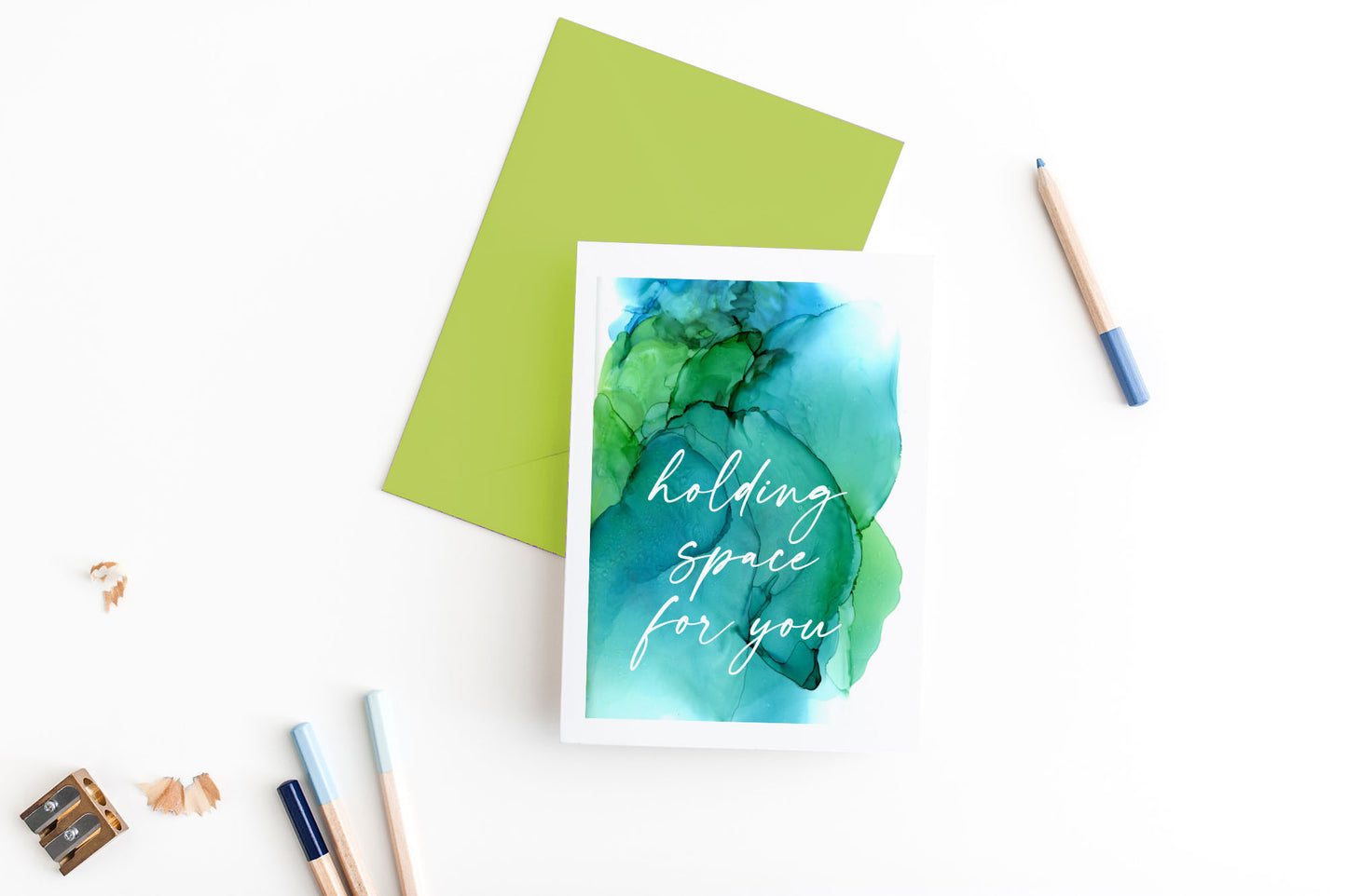 Greeting card, seen from above, with a colored, square flap envelope, pencils, pencil sharpener and shavings on a white background.