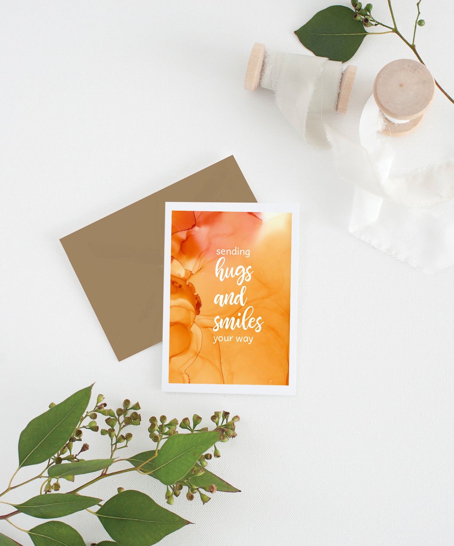 Greeting card with colorful envelope laying on a white surface with green leaves and cotton ribbon wound on wooden spools.