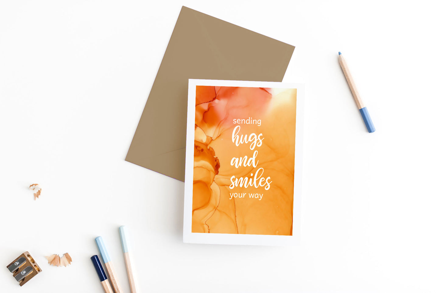 Greeting card, seen from above, with a colored, square flap envelope, pencils, pencil sharpener and shavings on a white background.