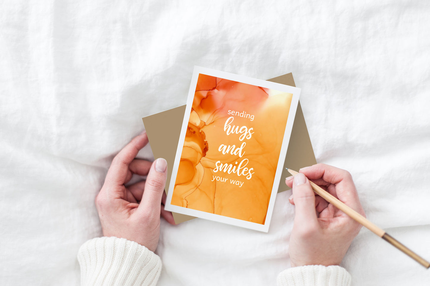 Greeting card and colorful envelope, seen from above, held by two hands along with a pencil, ready to write, against a white comforter.