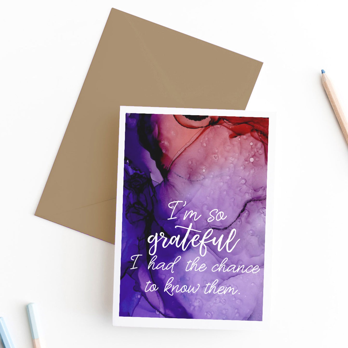Zoomed in view of greeting card, seen from above, with a kraft, square flap envelope with pencils on a white background.