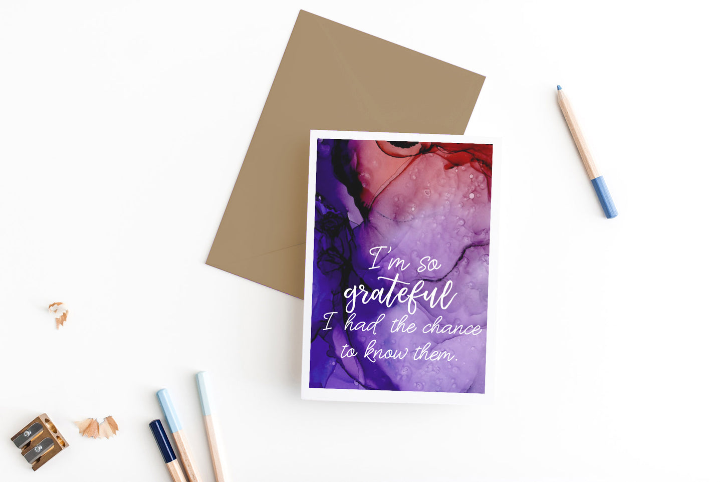 Greeting card, seen from above, with a kraft, square flap envelope, pencils, pencil sharpener and shavings on a white background.