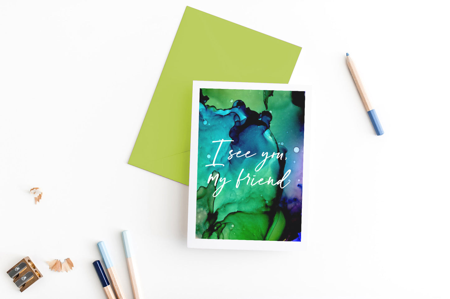 Greeting card, seen from above, with a colored, square flap envelope, pencils, pencil sharpener and shavings on a white background.