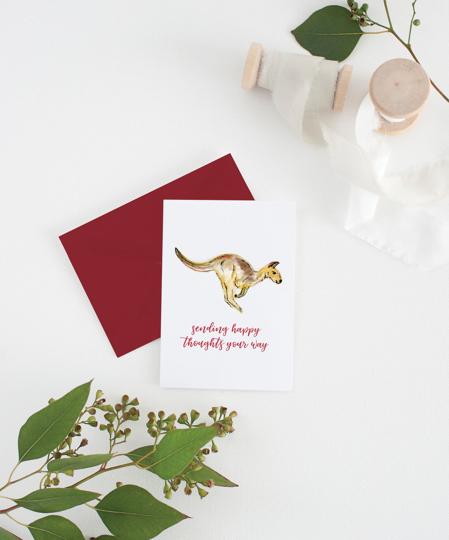 Greeting card with colorful envelope laying on a white surface with green leaves and cotton ribbon wound on wooden spools.
