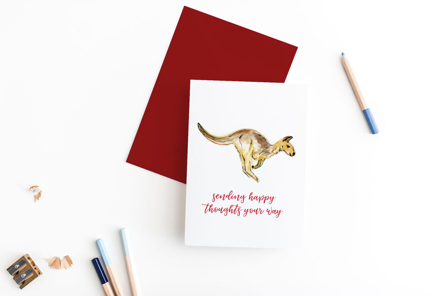 Greeting card, seen from above, with a colored, square flap envelope, pencils, pencil sharpener and shavings on a white background.