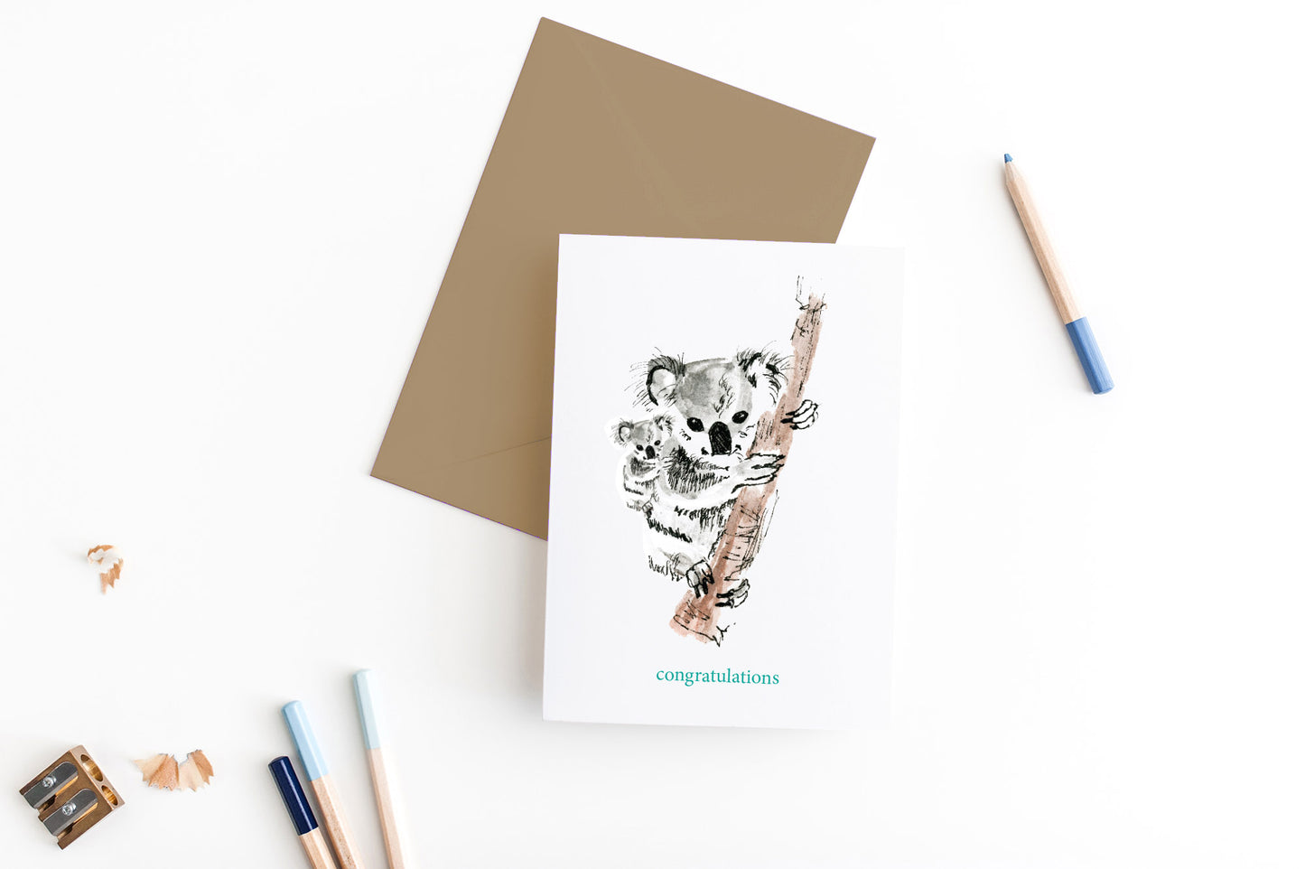 Greeting card, seen from above, with a kraft, square flap envelope, pencils, pencil sharpener and shavings on a white background.