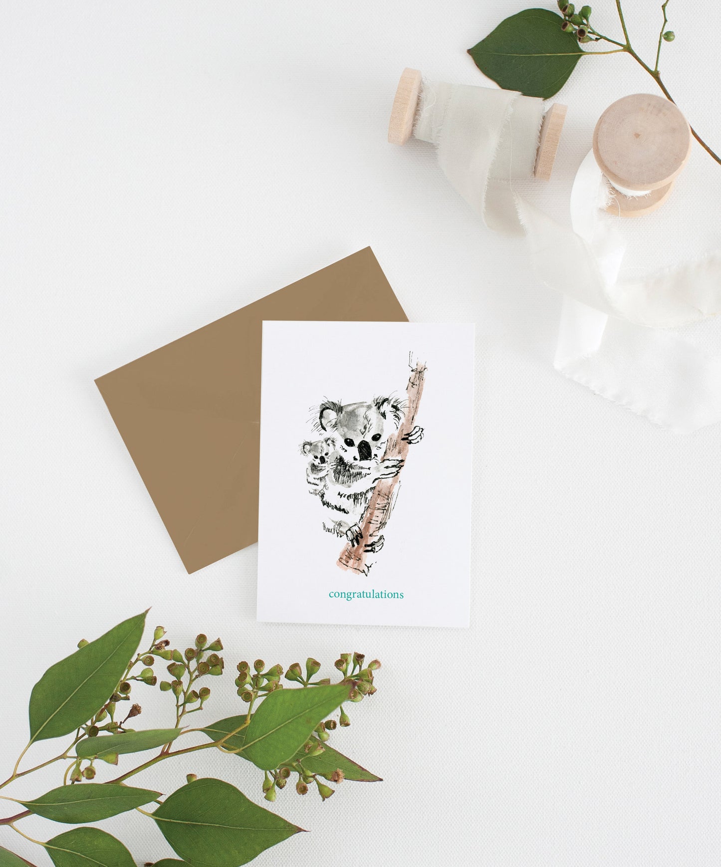 Greeting card with kraft envelope laying on a white surface with green leaves and cotton ribbon wound on wooden spools.