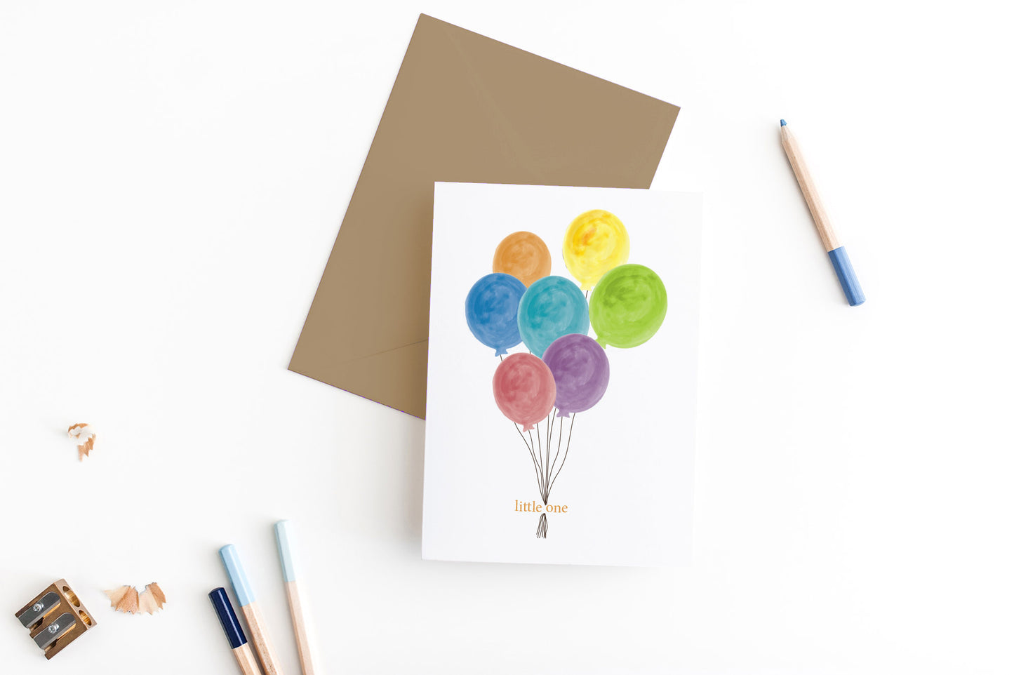 Greeting card, seen from above, with a kraft, square flap envelope, pencils, pencil sharpener and shavings on a white background.