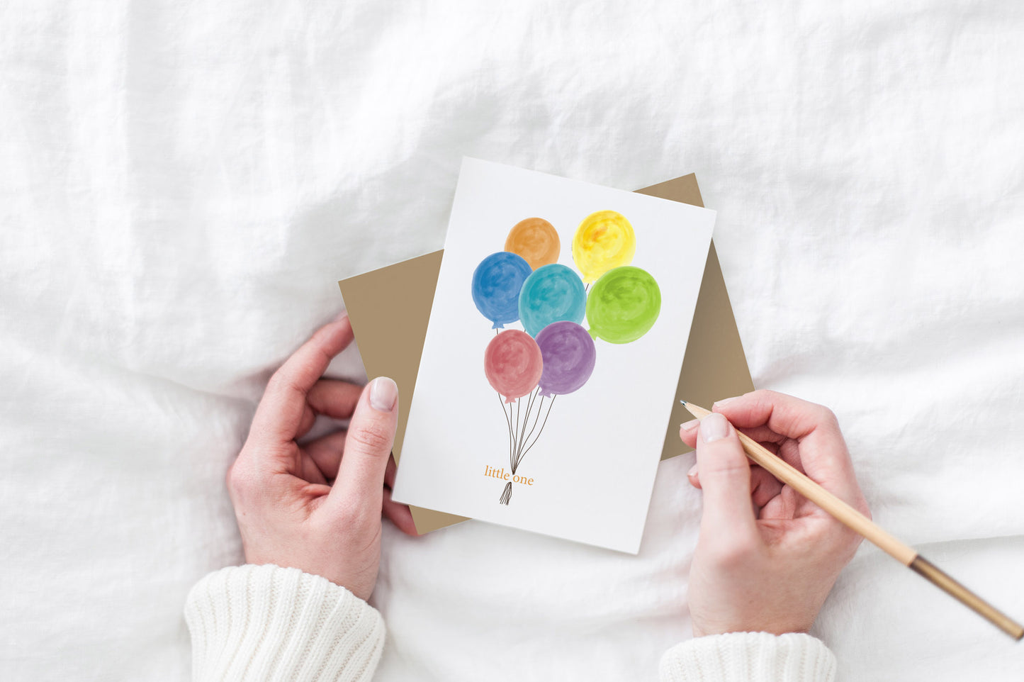 Greeting card and kraft envelope, seen from above, held by two hands along with a pencil, ready to write, against a white comforter.
