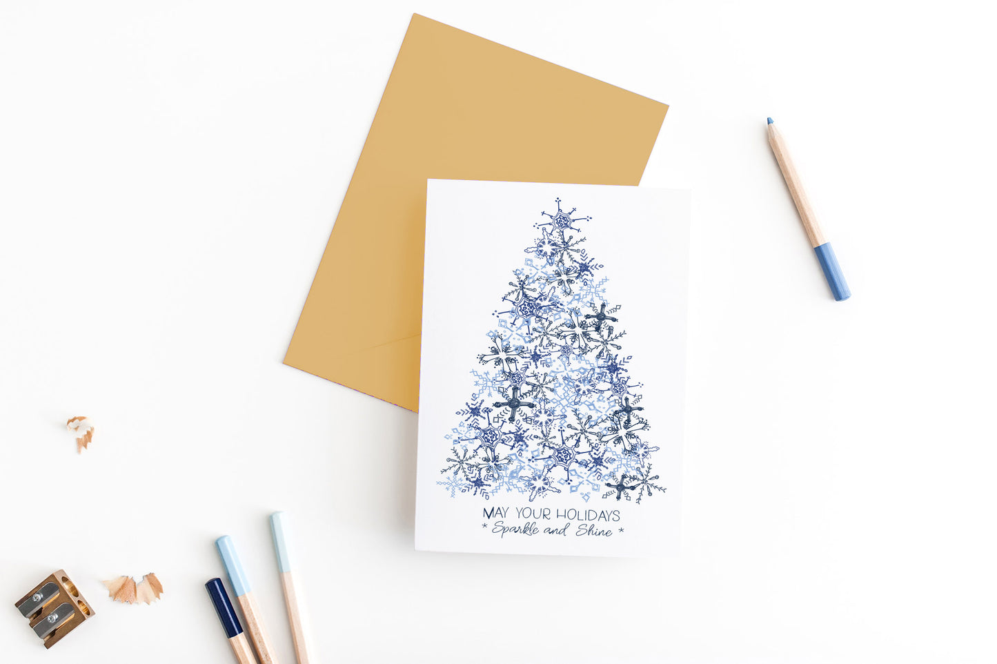 Greeting card, seen from above, with a colored, square flap envelope, pencils, pencil sharpener and shavings on a white background.