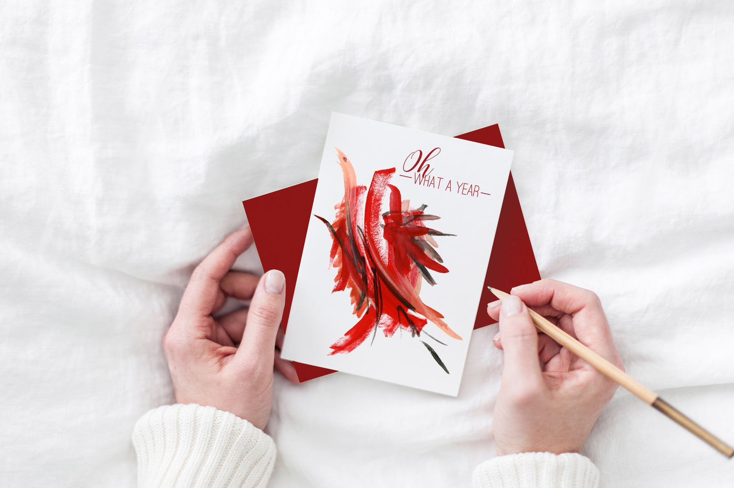 Greeting card and colorful envelope, seen from above, held by two hands along with a pencil, ready to write, against a white comforter.