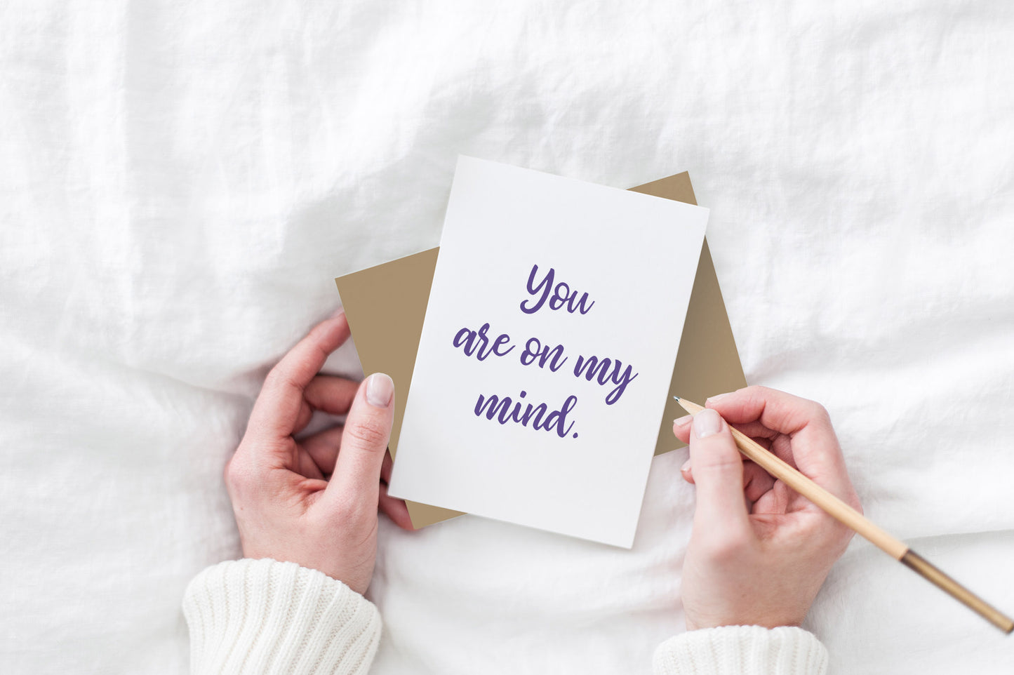 Greeting card and colorful envelope, seen from above, held by two hands along with a pencil, ready to write, against a white comforter.
