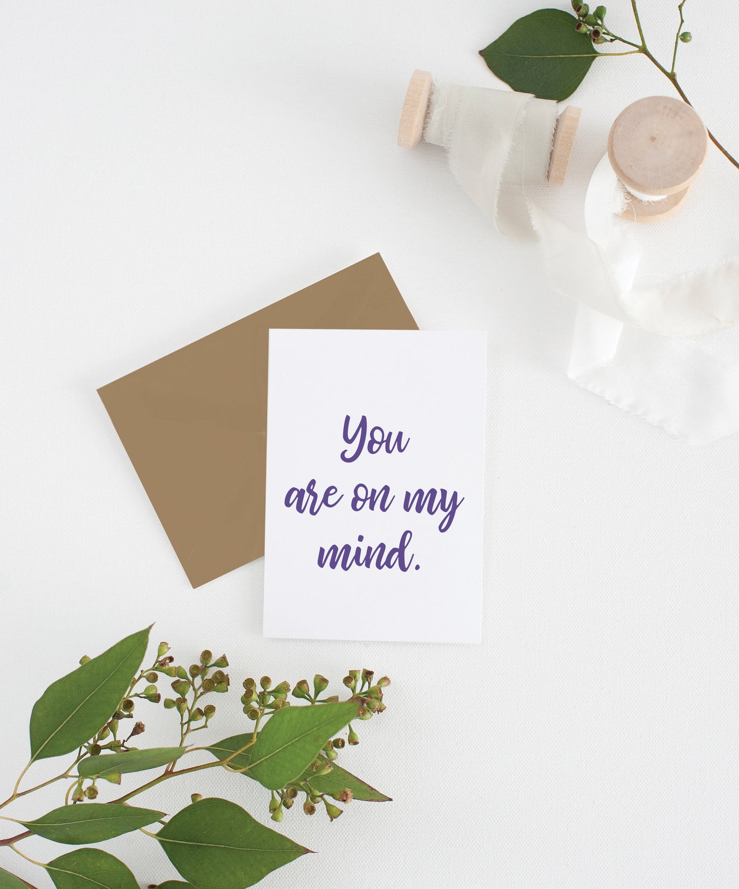 Greeting card with colorful envelope laying on a white surface with green leaves and cotton ribbon wound on wooden spools.