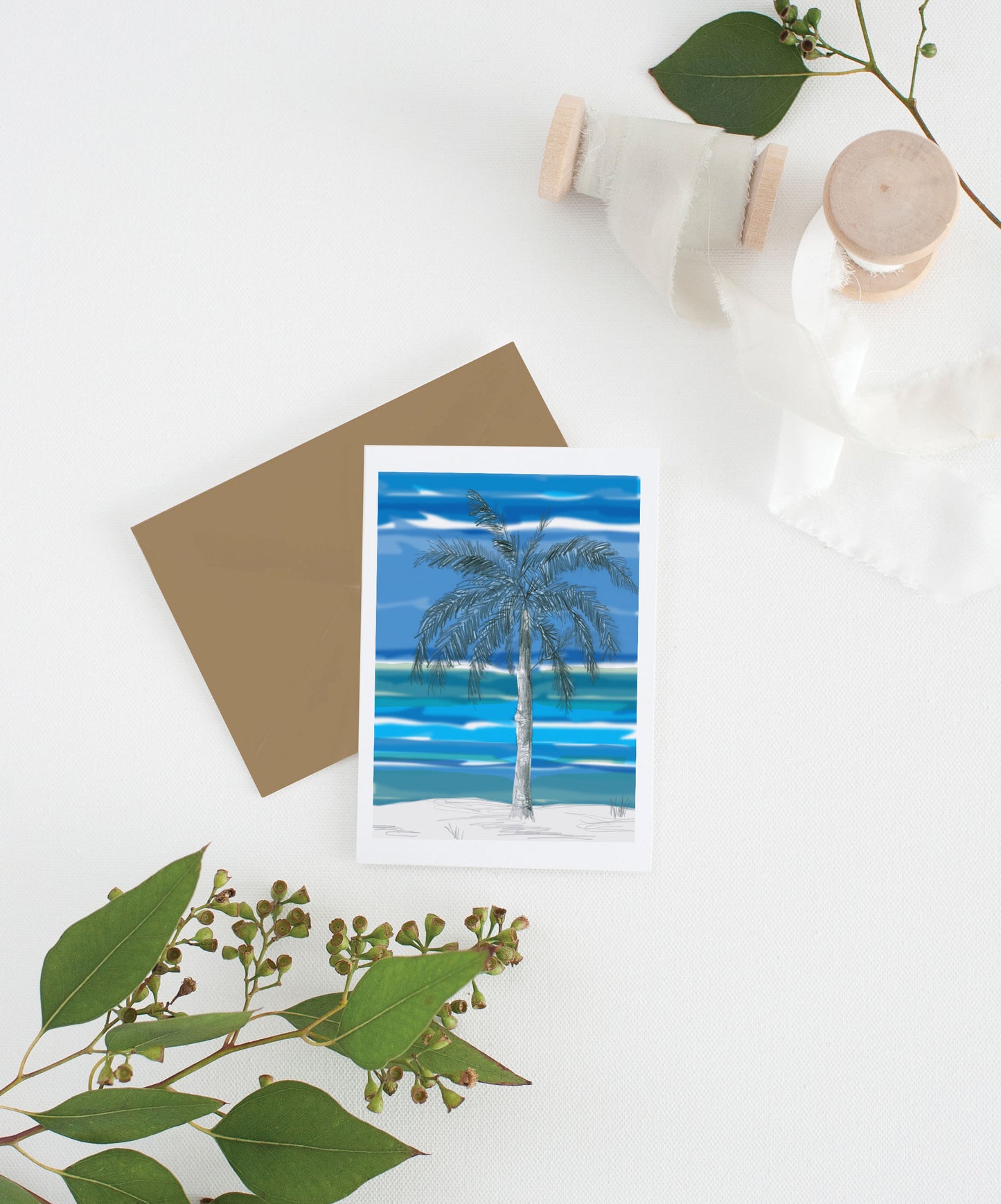 Greeting card with kraft envelope laying on a white surface with green leaves and cotton ribbon wound on wooden spools.