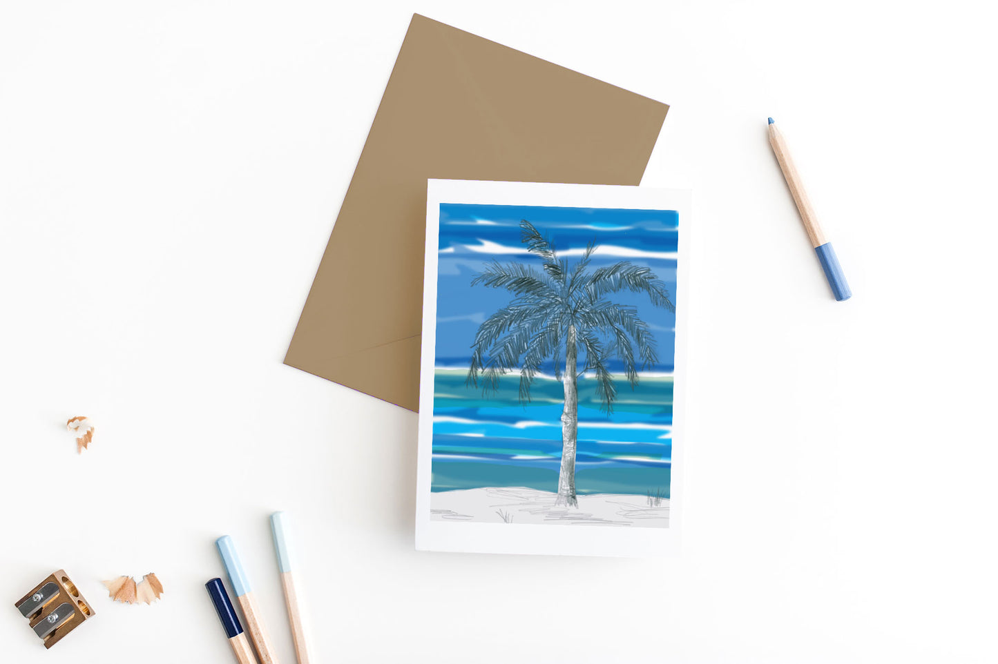 Greeting card, seen from above, with a kraft, square flap envelope, pencils, pencil sharpener and shavings on a white background.
