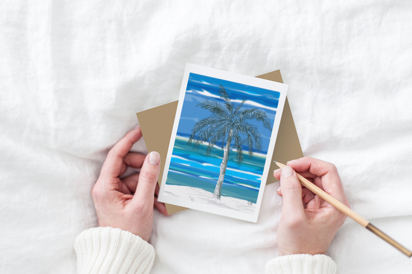 Greeting card and kraft envelope, seen from above, held by two hands along with a pencil, ready to write, against a white comforter.