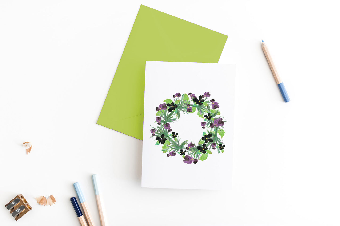 Greeting card, seen from above, with a colored, square flap envelope, pencils, pencil sharpener and shavings on a white background.