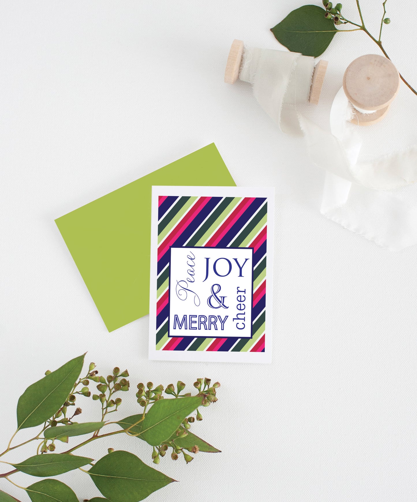 Greeting card with colorful envelope laying on a white surface with green leaves and cotton ribbon wound on wooden spools.