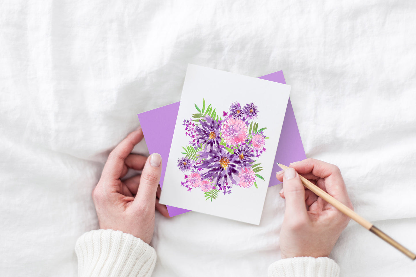 Greeting card and colorful envelope, seen from above, held by two hands along with a pencil, ready to write, against a white comforter.