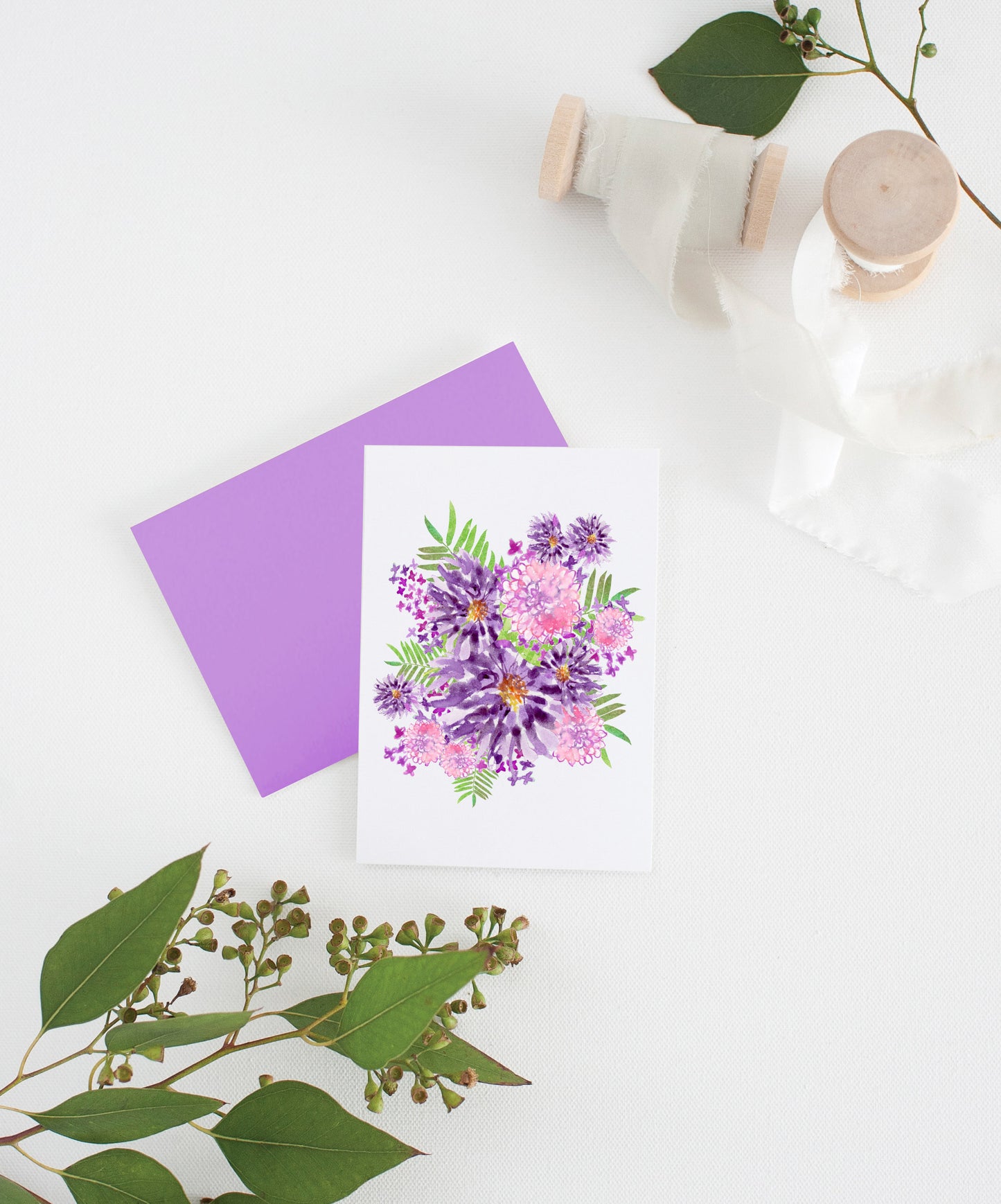 Greeting card with colorful envelope laying on a white surface with green leaves and cotton ribbon wound on wooden spools.