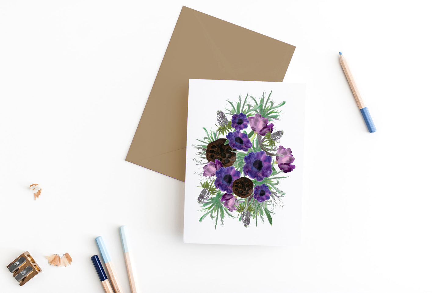 Greeting card, seen from above, with a kraft, square flap envelope, pencils, pencil sharpener and shavings on a white background.