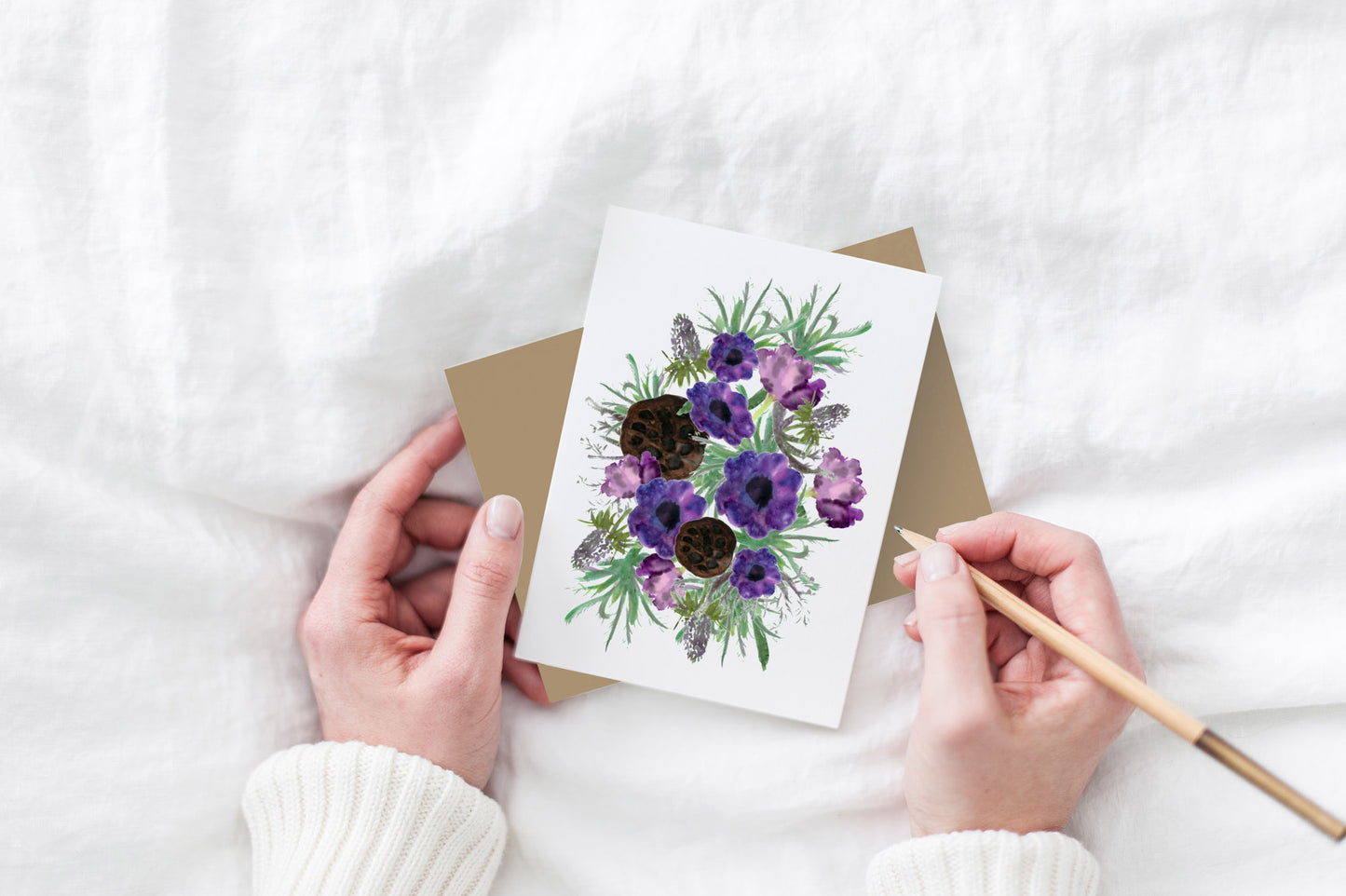 Greeting card and kraft envelope, seen from above, held by two hands along with a pencil, ready to write, against a white comforter.