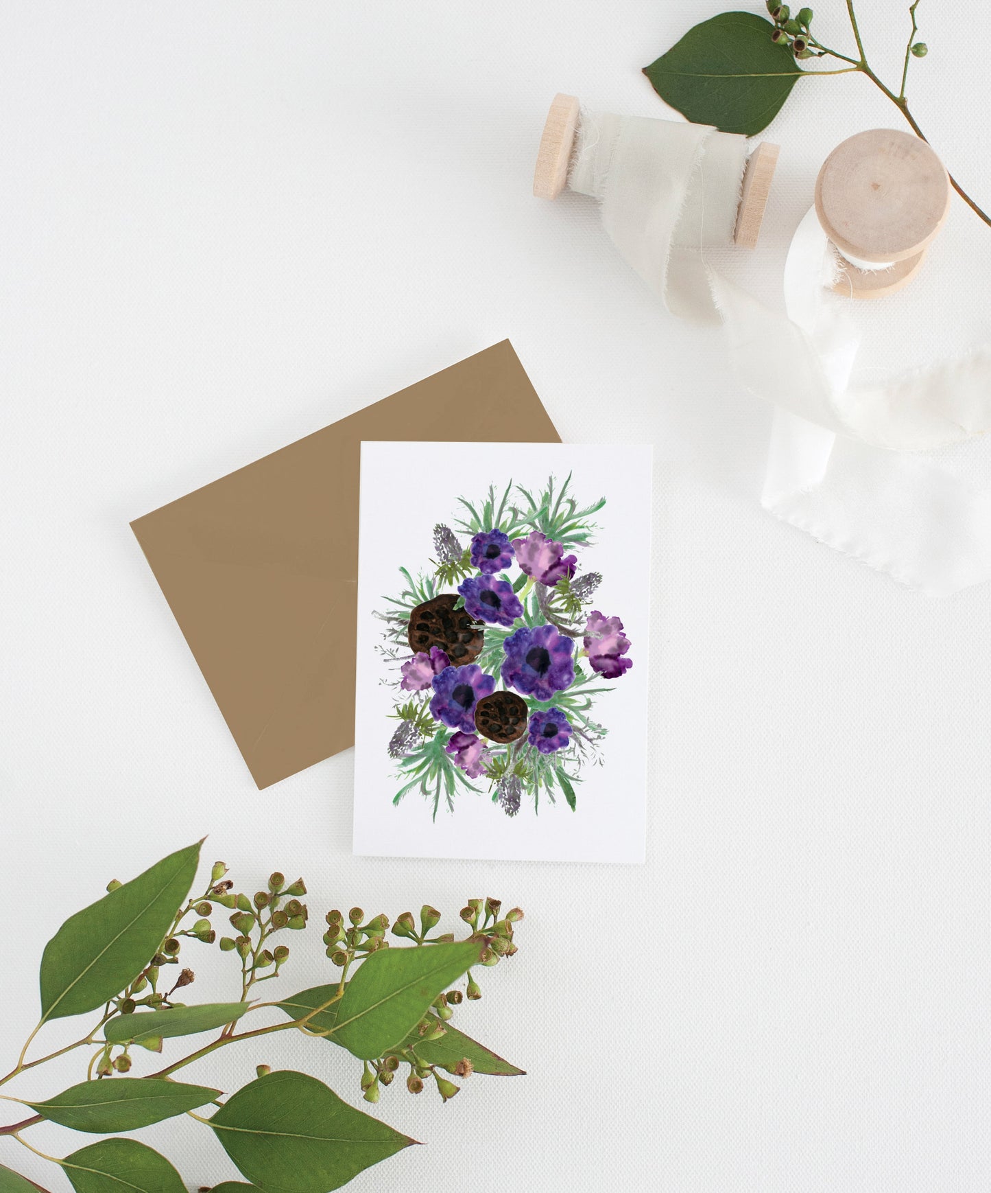 Greeting card with kraft envelope laying on a white surface with green leaves and cotton ribbon wound on wooden spools.