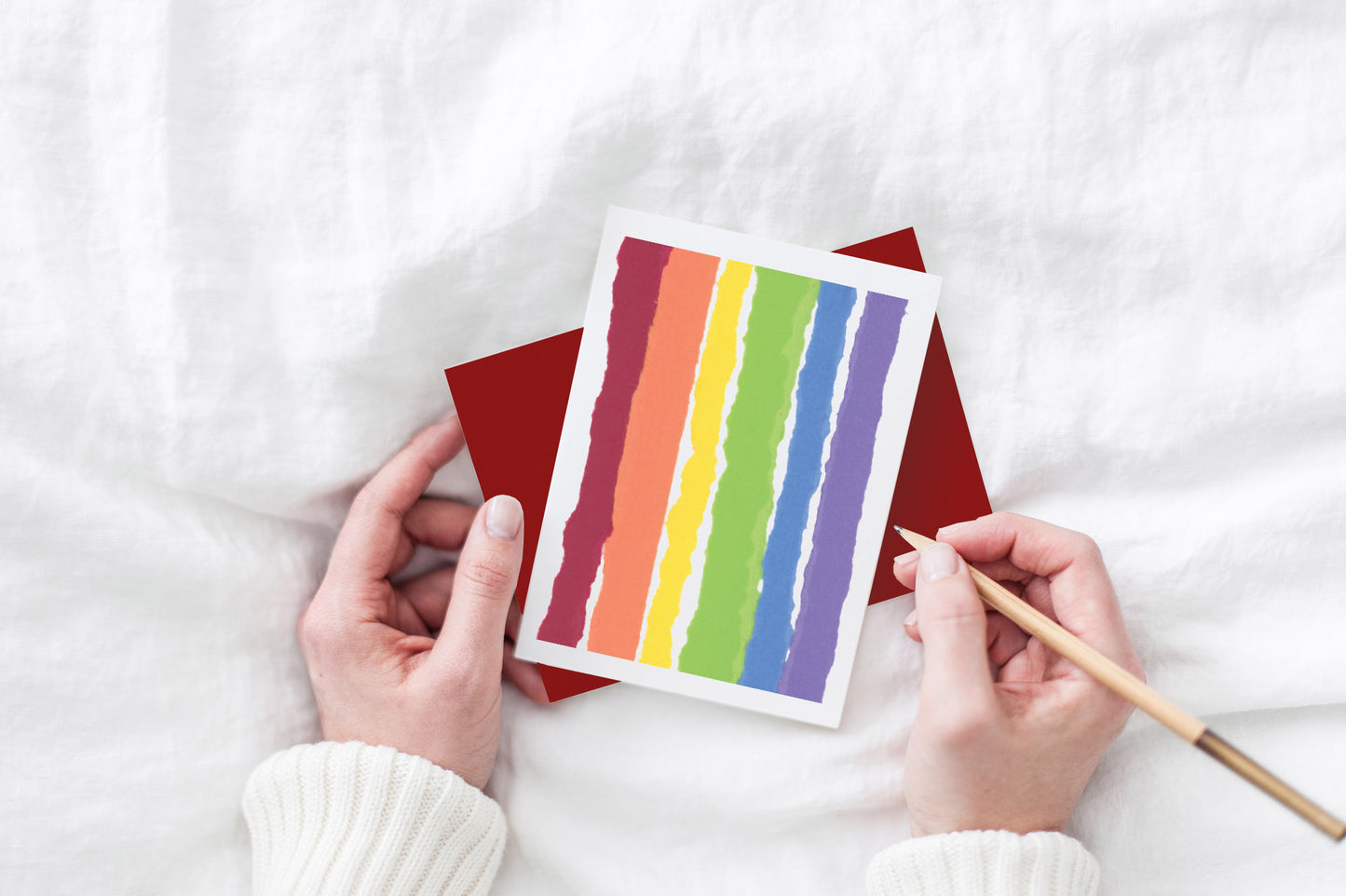 Greeting card and colorful envelope, seen from above, held by two hands along with a pencil, ready to write, against a white comforter.