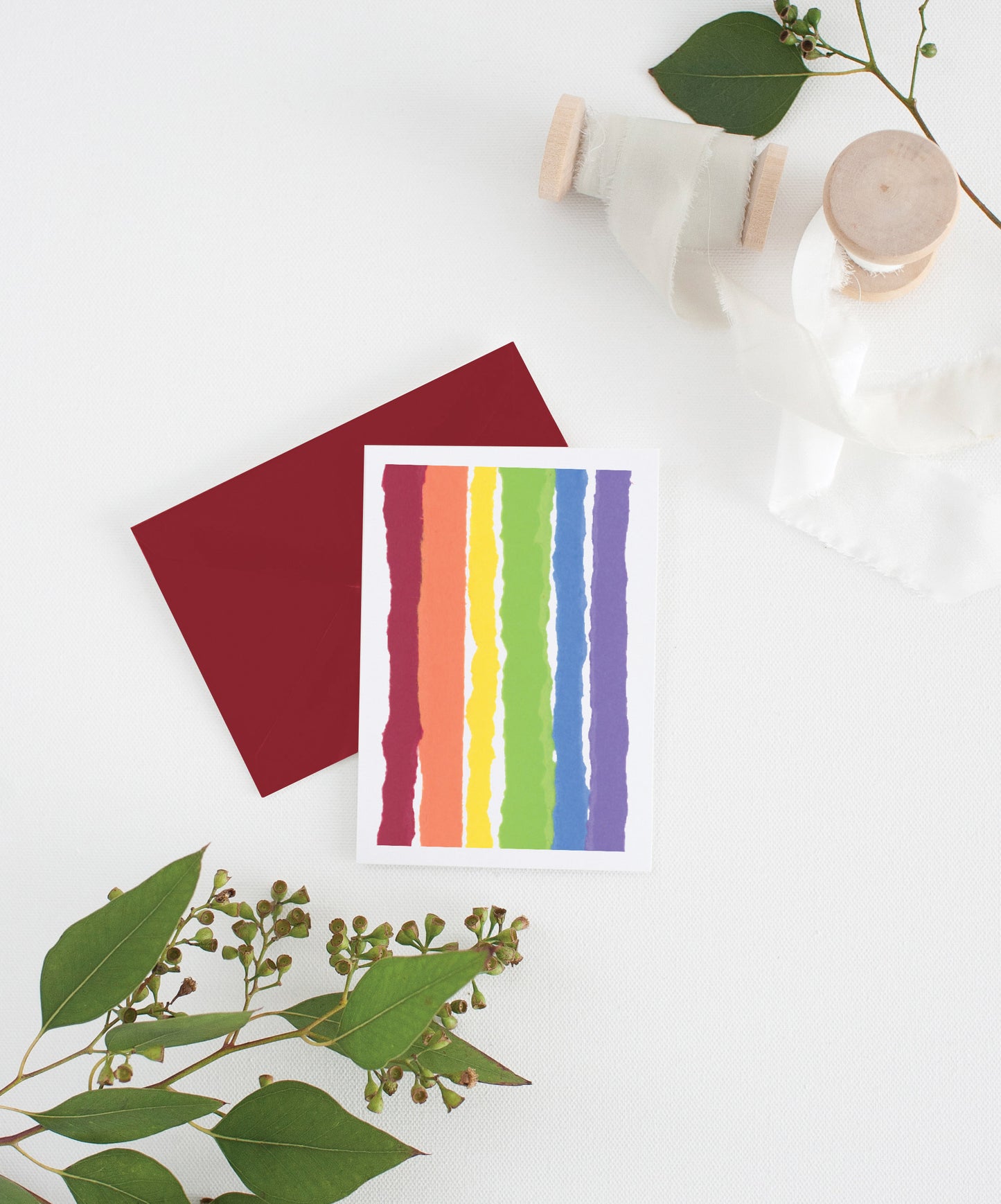 Greeting card with colorful envelope laying on a white surface with green leaves and cotton ribbon wound on wooden spools.