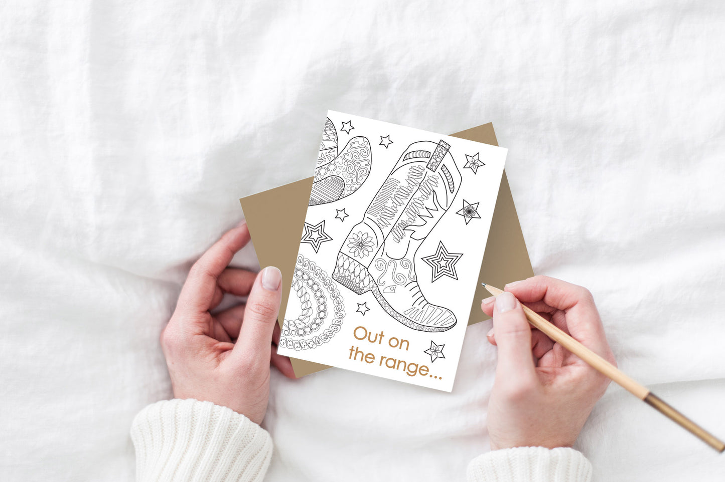 Greeting card and kraft envelope, seen from above, held by two hands along with a pencil, ready to write, against a white comforter.