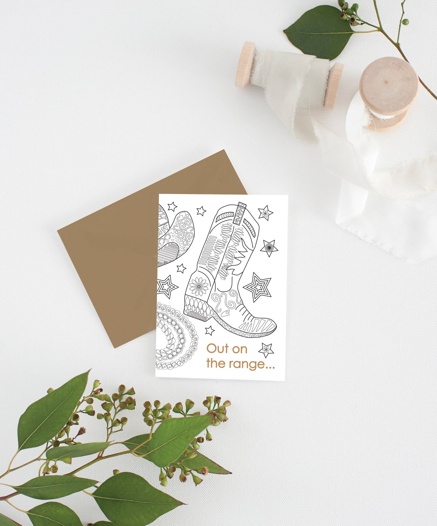 Greeting card with kraft envelope laying on a white surface with green leaves and cotton ribbon wound on wooden spools.