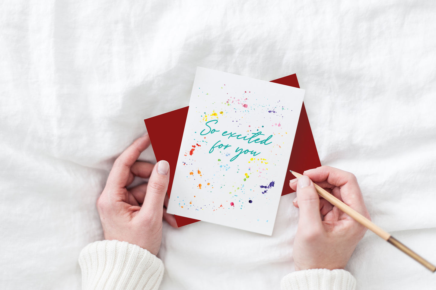 Greeting card and colorful envelope, seen from above, held by two hands along with a pencil, ready to write, against a white comforter.