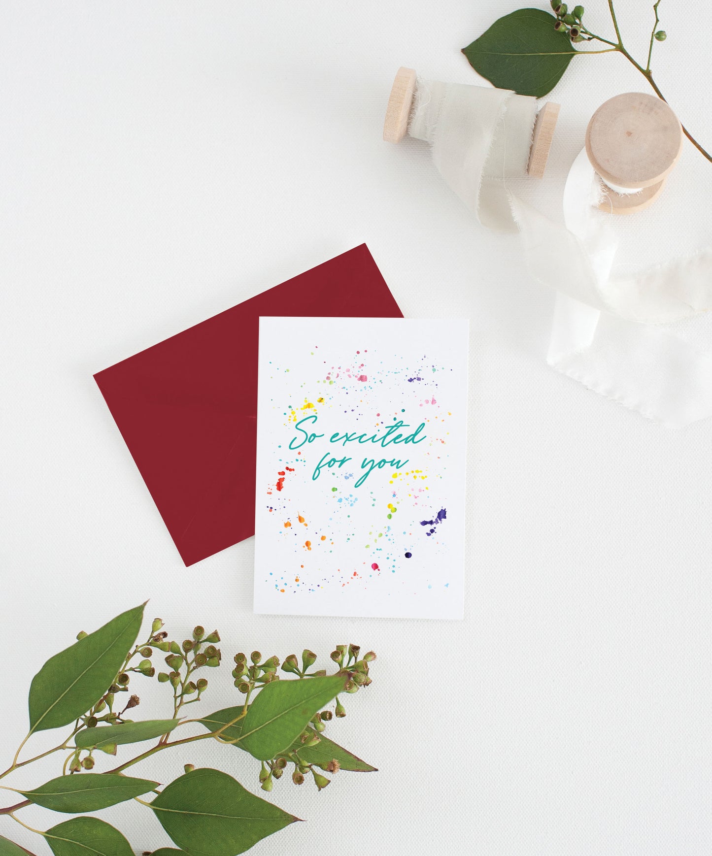 Greeting card with colorful envelope laying on a white surface with green leaves and cotton ribbon wound on wooden spools.