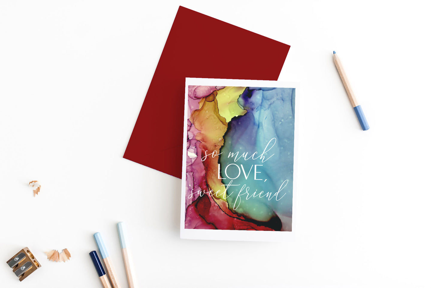 Greeting card, seen from above, with a colored, square flap envelope, pencils, pencil sharpener and shavings on a white background.