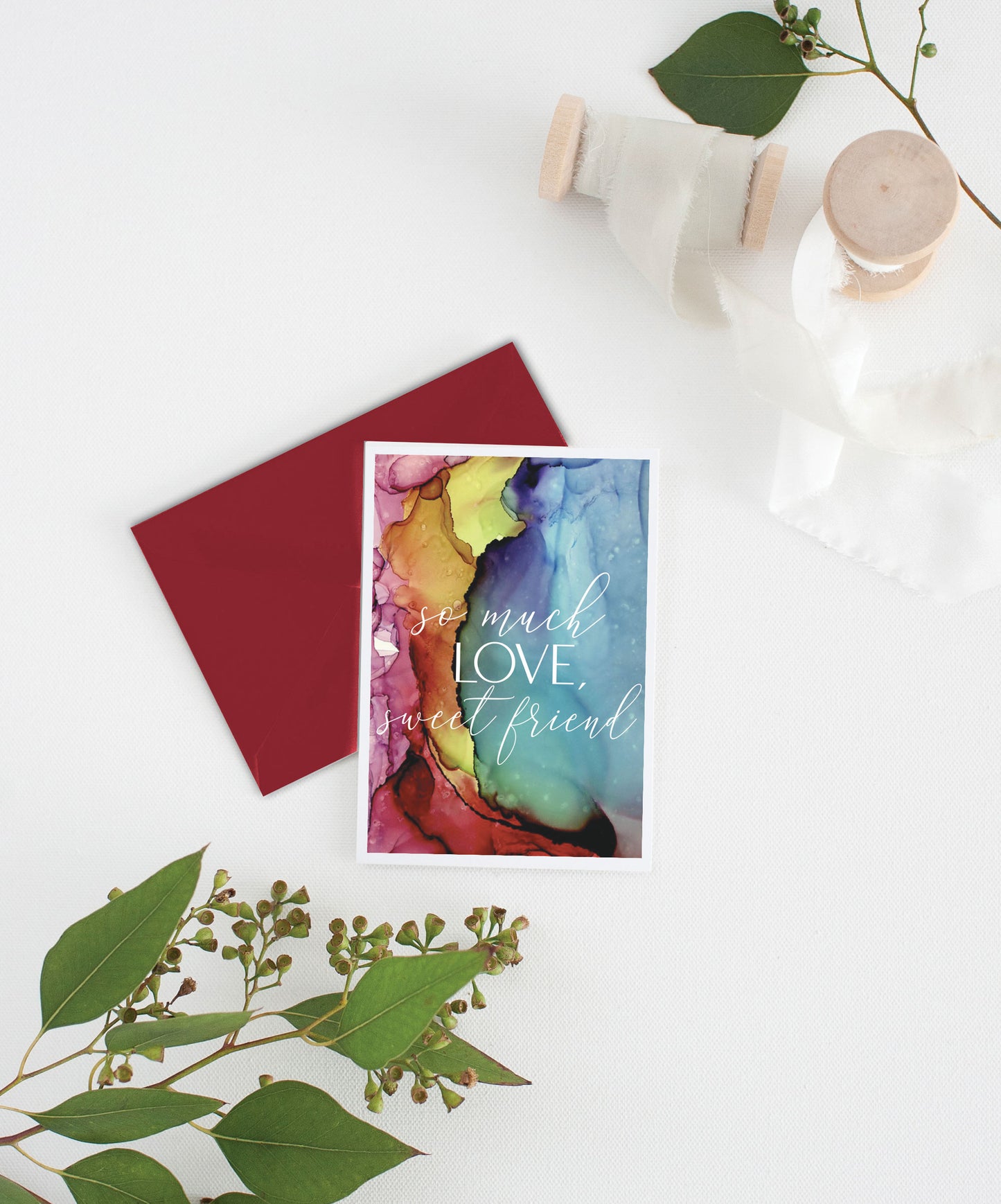 Greeting card with colorful envelope laying on a white surface with green leaves and cotton ribbon wound on wooden spools.