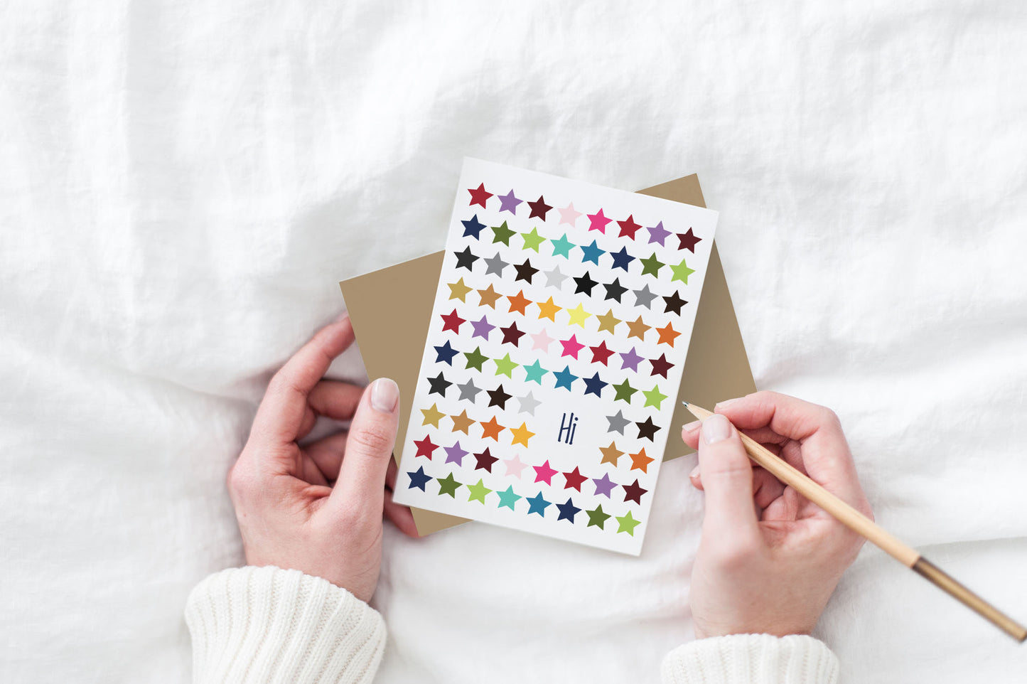 Greeting card and colorful envelope, seen from above, held by two hands along with a pencil, ready to write, against a white comforter.
