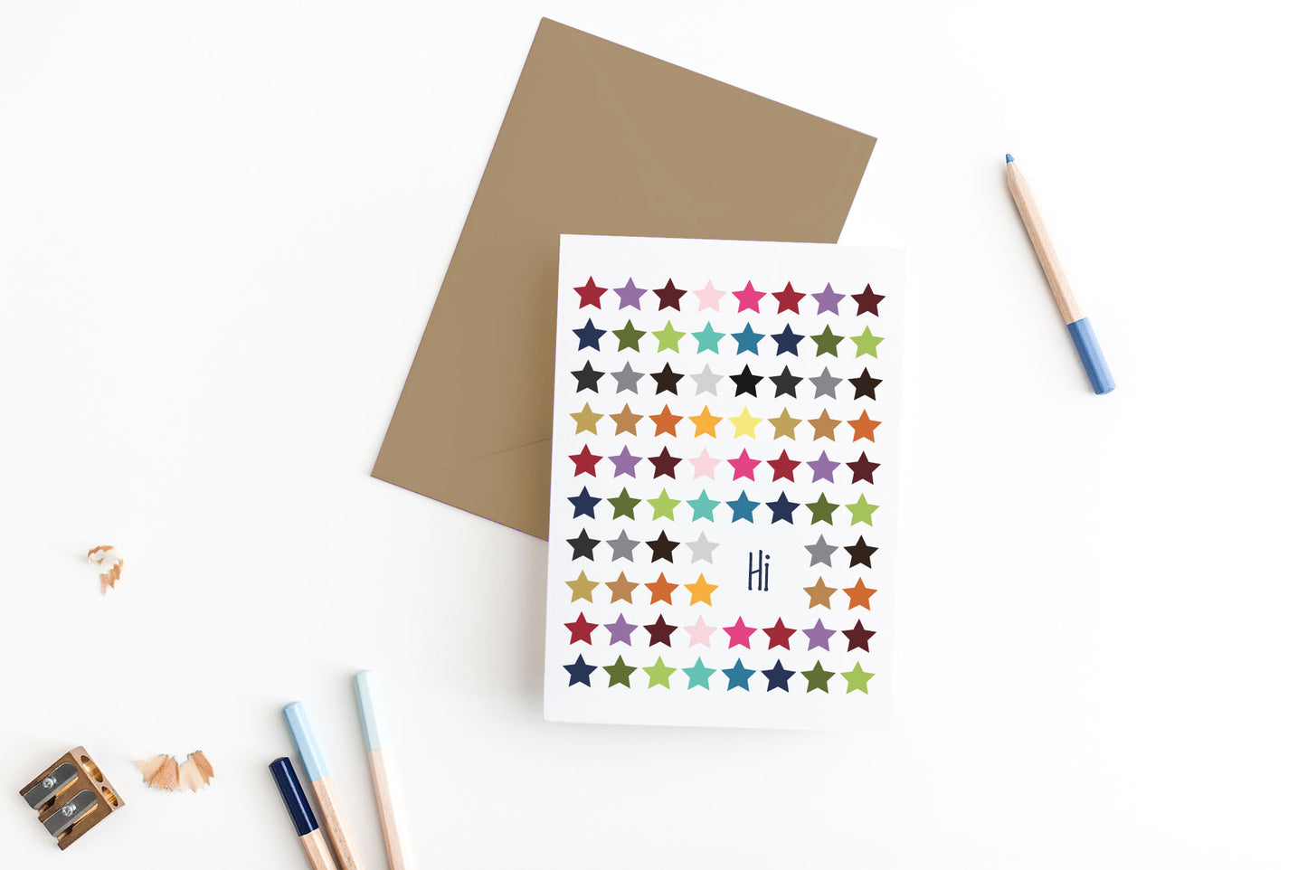 Greeting card, seen from above, with a kraft, square flap envelope, pencils, pencil sharpener and shavings on a white background.