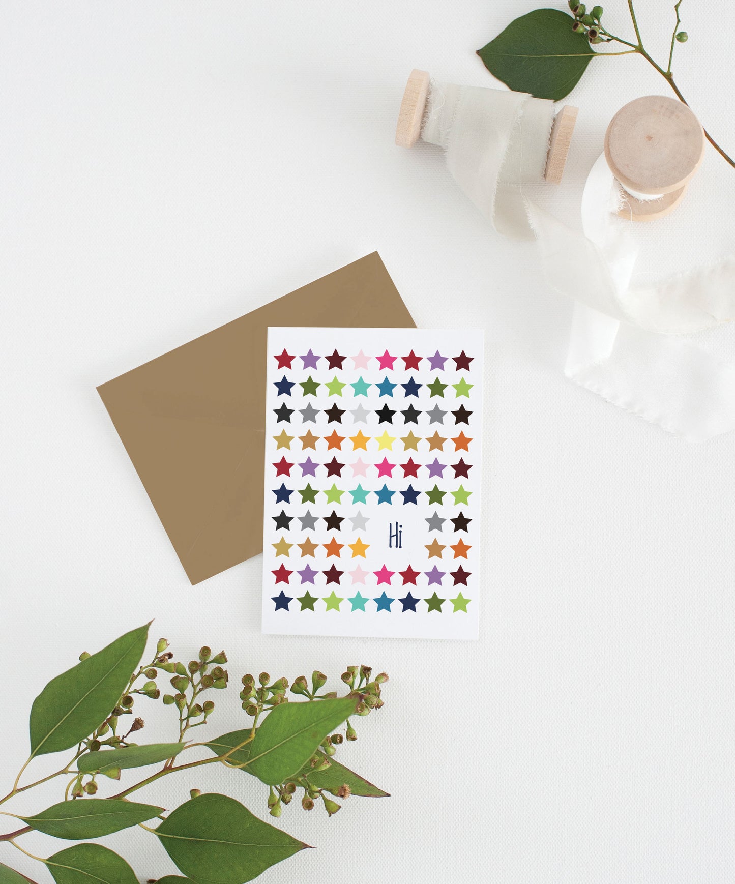 Greeting card with colorful envelope laying on a white surface with green leaves and cotton ribbon wound on wooden spools.