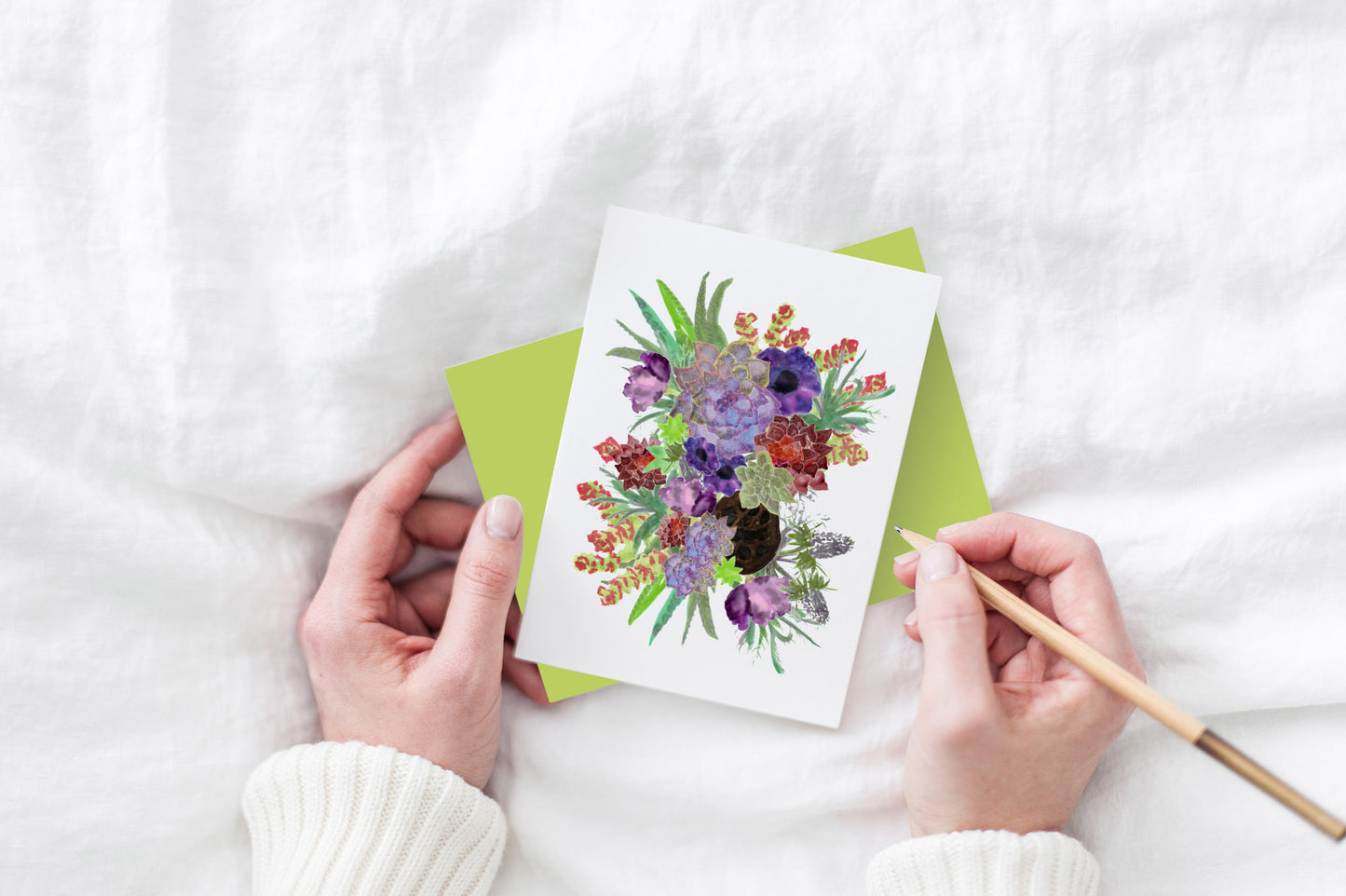 Greeting card and colorful envelope, seen from above, held by two hands along with a pencil, ready to write, against a white comforter.