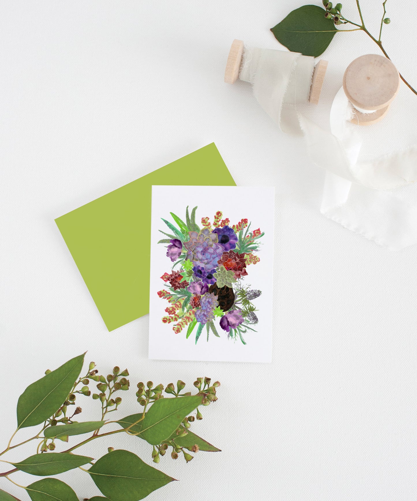 Greeting card with colorful envelope laying on a white surface with green leaves and cotton ribbon wound on wooden spools.