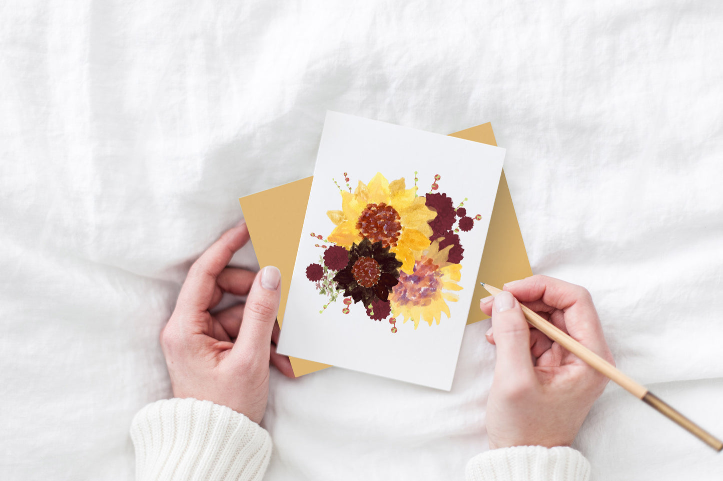 Greeting card and colorful envelope, seen from above, held by two hands along with a pencil, ready to write, against a white comforter.
